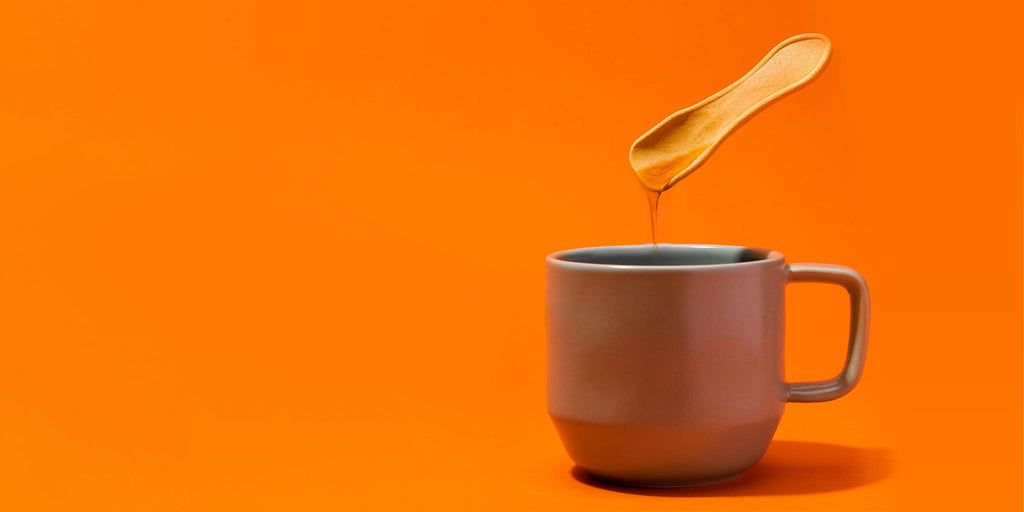 A photo of a small edible spoon drizzling honey into a cup of tea. The tea mug is on an orange background.